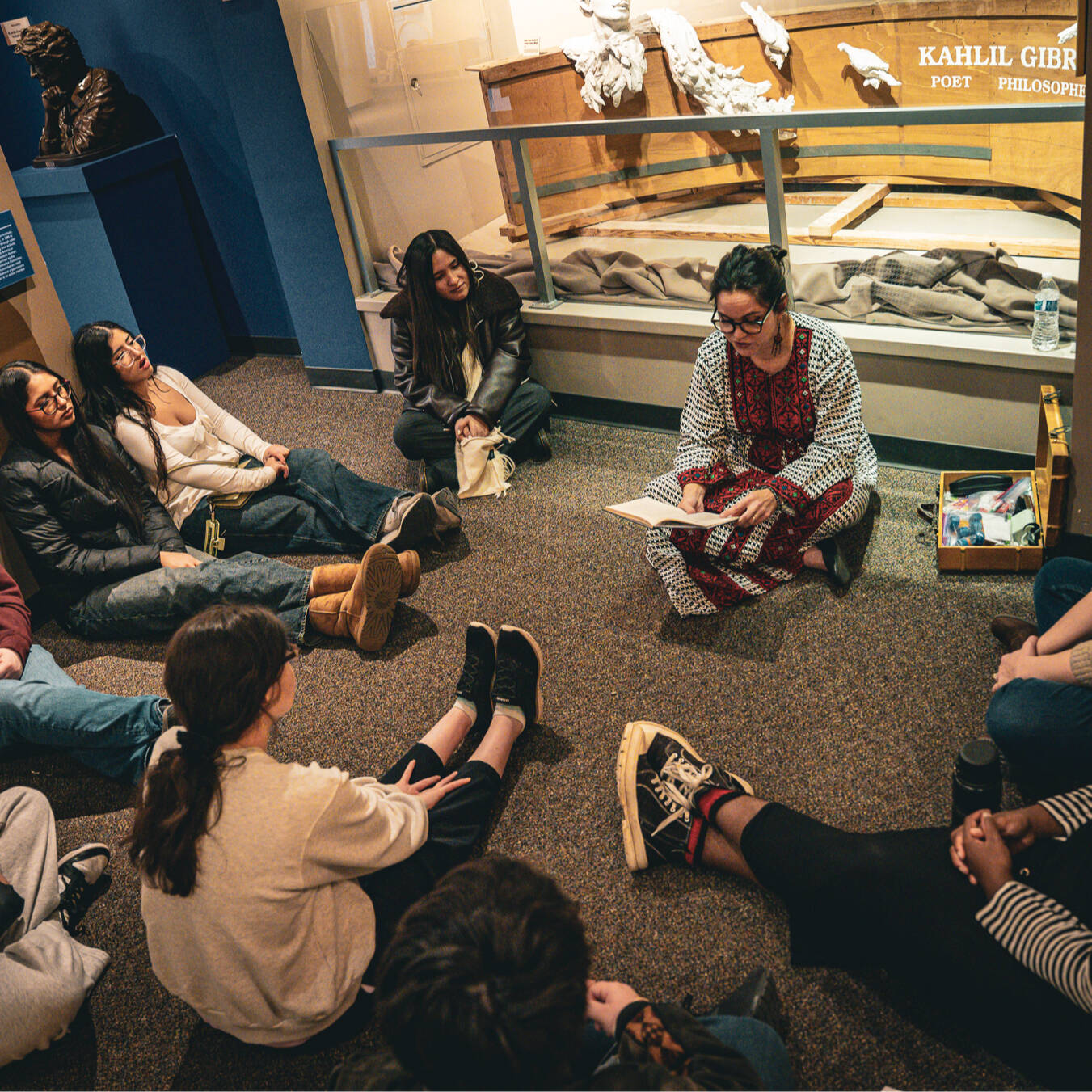 A group of students sitting in a circle on the floor listening to a female tour guide read from a book of poetry.